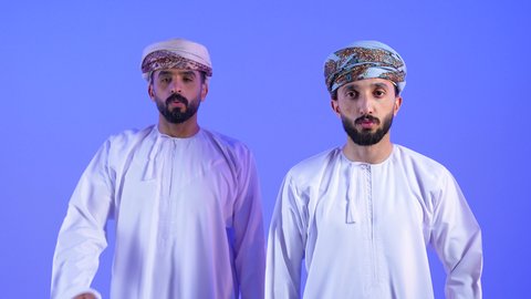 الانزعاج عند سماع خبر سيء ,الوقوع في مصيبة, النظر الى الكاميرا بتعابير التعجب, الصدمة و المفاجأة برؤية شيء ما، بورتريه لرجلان عربيان خليجيان عمانيان يرتديان الدشداشة و العمامه   يضعان يدهما على وجههم بتعابير الدهشة و الذهول, خلفية زرقاء