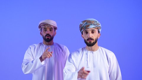 طلب السكوت والصمت, التوقف عن الكلام وخفض الصوت، الهدوء والسكون وعدم الإزعاج، بورتريه مقرب لرجلان عربيان خليجيان عمانيان يرتديان الدشداشة و العمامة يقفان باستقامة ويضعان أصبع السبابة على فمهما, خلفية زرقاء