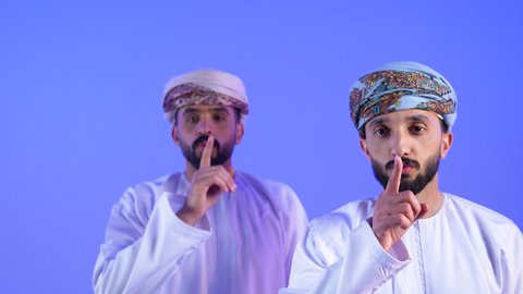 التوقف عن الكلام وخفض الصوت، الهدوء والسكون وعدم الإزعاج، بورتريه مقرب لرجلان  عربيان خليجيان عمانيان يرتديان الدشداشة و العمامة يقفان باستقامة ويضعان أصبع السبابة على فمهما، طلب السكوت والصمت, خلفية زرقاء