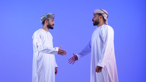 إيماءات السرور والبشاشة والترحيب، عقد صفقة واتفاقات، إلقاء التحية والسلام, بورتريه لشابين عربيين خليجيين عمانيين مبتسميين يرتديان الدشداشة و العمامة يقومان بمصافحة بعضهم, مفهوم الاستقبال والترحيب ,خلفية زرقاء