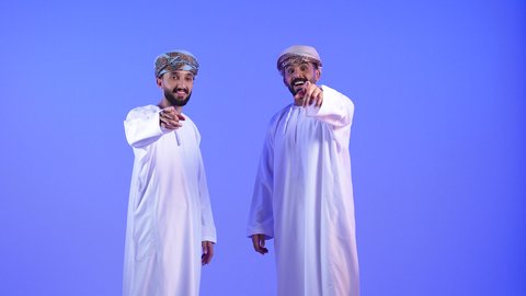 تعابير التأكد والتحديد, بورتريه لشابان عربيان خليجيان عمانيان يرتديان الدشداشة و العمامة ينظانر الى الكاميرا يشيران بأصابعهما السبابة الى شيء ما بإيماءات السرور، تعابير البهجة والفرح, خلفية زرقاء