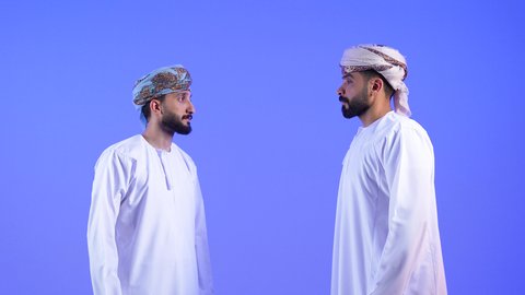 بناء علاقات و تكوين صداقات جديدة، الترحيب و السلام و إلقاء التحية، بورتريه لشابين عربين خليجين عمانين يرتديان الدشداشة و العمامة يقومان بتوجيه قبضة يده مقابل يد شخص آخر، ايماءات وحركات يدين تدل على التشجيع، خلفية زرقاء