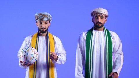 ارتداء اوشحة تشجيعية للفريق المفضل, تعابير الانزعاج لخسارة المنتخب الوطني، الشغف في متابعة الأندية الرياضية منذ الصغر، تشجيع الفريق العماني، بورتريه لرجل عربي خليجي عماني يرتدي الدشداشة و العمامة تظهر عليه ايماءات الخيبة برفقه صديقة, مشاهدة الفريق المفضل بتعابير التفاعل والاندماج وامساك كرة القدم، خلفية زرقاء