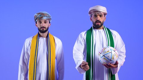 تعابير الانزعاج لخسارة المنتخب الوطني, ارتداء اوشحة تشجيعية للفريق المفضل، الشغف في متابعة الأندية الرياضية منذ الصغر، تشجيع الفريق العماني، بورتريه لرجل عربي خليجي عماني يرتدي الدشداشة و العمامة تظهر عليه ايماءات الخيبة برفقه صديقة, مشاهدة الفريق المفضل بتعابير التفاعل والاندماج وامساك كرة القدم، خلفية زرقاء