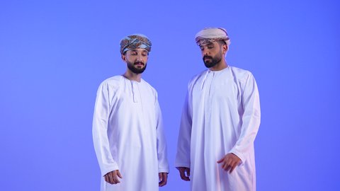 النظر الى الكاميرا بتعابير السرور، تقوية وتعزيز الروابط العائلية، مفهوم الصداقة والأخوة، المحبة والمودة بين الأخوات، بورتريه لشابين عربيين خليجيين عمانيين مبتسمين يرتديان الدشداشة و العمامة يرفعان يديهما ويشكلان قلب حب, خلفية زرقاء