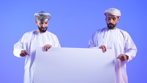 Empty square white board, marketing offers and advertisements, portrait of two Omani Gulf Arab men wearing dishdasha and turban holding a white board in their hands and pointing to it, blue background
