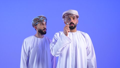 الصدمة والذهول لرؤية شيء ما, مفهوم الفحص والاستكشاف، توضيح الصورة والرؤية عن قرب، بورتريه لرجل عربي خليجي عماني يرتدي الدشداشة والعمامة يحمل بيده عدسة مكبرة يضعها على عينه بإيماءات التركيز والتمعن برفقه صديقة , خلقية زرقاء