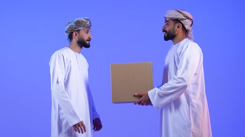 إستلام المشتريات و تسليم البضائع للزبائن, توصيل الطلبيات بالوقت المحدد و بشكل سليم, بورتريه لرجلان عربيان خليجيان عمانيان يرتديان الدشداشة و العمامة يحملان بيدهما صندوق كرتوني مغلق, النظر الى الكاميرا بتعابير السرور, خلفية زرقاء
