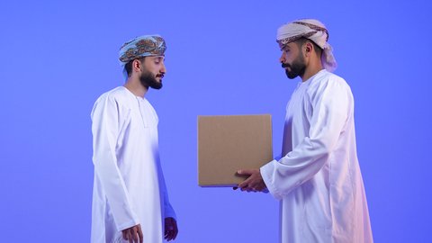 توصيل الطلبيات بالوقت المحدد و بشكل سليم, بورتريه لرجلان عربيان خليجيان عمانيان يرتديان الدشداشة و العمامة يحملان بيدهما صندوق كرتوني مغلق, إستلام المشتريات و تسليم البضائع للزبائن, النظر الى الكاميرا بتعابير السرور, خلفية زرقاء