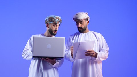 مفهوم استخدام التكنولوجيا الحديثة في أداء المهام والعمل عن بعد، التعاون والتشارك بين الزملاء في اختيار خطة العمل، بورتريه لرجلان عربيان خليجيان عمانيان يرتديان الزي العماني التقليدي يقومان بالعمل على جهاز الاب توب المحمول في متابعه سير العمل، خلفية زرقاء