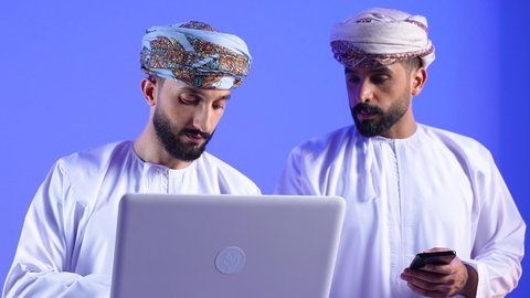 التواصل مع الأهل والأصدقاء، تصفح مواقع التواصل الاجتماعي عبر الجوال، مشاهدة الأفلام والمسلسلات الممتعة، استخدام التكنولوجيا و التقنيات الحديثة، رجلان عربيان خليجيان عمانيان يرتديان الدشداشة و العمامه يقومان باستخدام الهاتف و الحاسب المحمول ، خلفية زرقاء