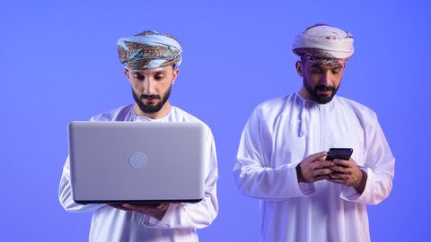 تصفح مواقع التواصل الاجتماعي عبر الجوال، مشاهدة الأفلام والمسلسلات الممتعة، التواصل مع الأهل والأصدقاء، استخدام التكنولوجيا و التقنيات الحديثة، رجلان عربيان خليجيان عمانيان يرتديان الدشداشة و العمامه يقومان باستخدام الهاتف و الحاسب المحمول ، خلفية زرقاء