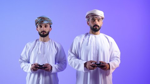 Modern advanced technologies, the concept of entertainment, amusement, winning and triumph, a portrait of two Omani Gulf Arab men wearing a dishdasha and a turban holding a joystick, enjoying playing video games in their spare time, challenge, adventure and competition, blue background