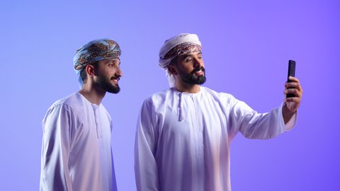 اجراء مكالمة فيديو عبر تطبيقات ومواقع السوشال ميديا، قضاء الوقت الممتع مع الاخوة، استخدام جهاز تقني حديث ومتطور، توثيق اللحظات السعيدة عبر الهاتف المحمول, بورتريه لشاب عربي خليجي عماني يرتدي الدشداشة و العمامة يقوم بالتقاط صورة السيلفي التذكارية مع صديقة عبر الجوال، خلفية زرقاء