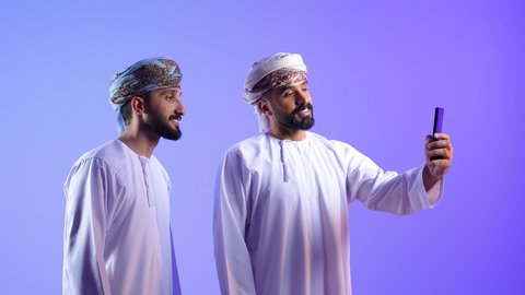 قضاء الوقت الممتع مع الاخوة، استخدام جهاز تقني حديث ومتطور، توثيق اللحظات السعيدة عبر الهاتف المحمول, بورتريه لشاب عربي خليجي عماني يرتدي الدشداشة و العمامة يقوم بالتقاط صورة السيلفي التذكارية مع صديقة عبر الجوال، خلفية زرقاء