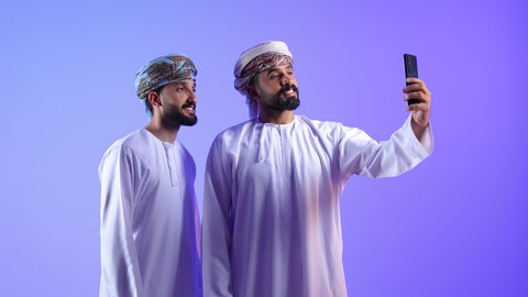 استخدام جهاز تقني حديث ومتطور، توثيق اللحظات السعيدة عبر الهاتف المحمول, قضاء الوقت الممتع مع الاخوة, بورتريه لشاب عربي خليجي عماني يرتدي الدشداشة و العمامة يقوم بالتقاط صورة السيلفي التذكارية مع صديقة عبر الجوال، خلفية زرقاء