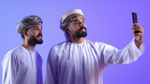 توثيق اللحظات السعيدة عبر الهاتف المحمول, استخدام جهاز تقني حديث ومتطور, قضاء الوقت الممتع مع الاخوة, بورتريه لشاب عربي خليجي عماني يرتدي الدشداشة و العمامة يقوم بالتقاط صورة السيلفي التذكارية مع صديقة عبر الجوال، خلفية زرقاء