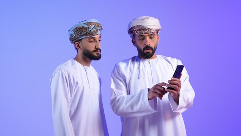 استخدام جهاز تقني حديث ومتطور, التسلية والترفيه في وقت الفراغ, بورتريه لشابين عربيين خليجيين عمانيين يرتديان الدشداشة و العمامة يقومان بصفح وسائل التواصل الاجتماعي من خلال الهاتف المحمول، خلفية زرقاء