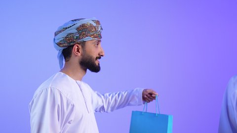 متعة التسوق في مولات ومتاجر السلطنة، تعابير السعادة للاستفادة من العروض و التخفيضات، اقتناء المستلزمات والهدايا, بورتريه لرجلين عربين خليجين عمانين يرتديان الدشداشة و العمامة يحملان بيدهما الأكياس الورقية، خلفية زرقاء
