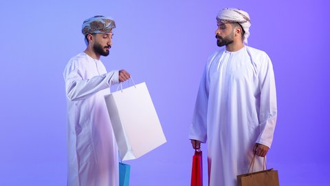 تعابير السعادة للاستفادة من العروض و التخفيضات، اقتناء المستلزمات والهدايا, بورتريه لرجلين عربين خليجين عمانين يرتديان الدشداشة و العمامة يحملان بيدهما الأكياس الورقية, متعة التسوق في مولات ومتاجر السلطنة، خلفية زرقاء