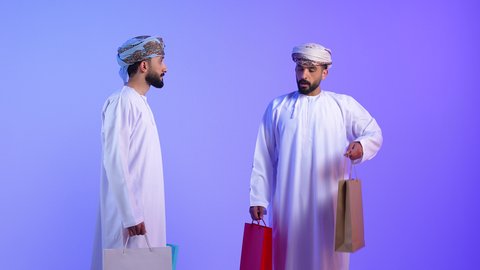 اقتناء المستلزمات والهدايا, تعابير السعادة للاستفادة من العروض و التخفيضات, بورتريه لرجلين عربين خليجين عمانين يرتديان الدشداشة و العمامة يحملان بيدهما الأكياس الورقية, متعة التسوق في مولات ومتاجر السلطنة، خلفية زرقاء