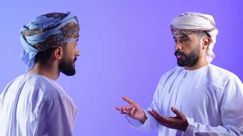 Having a good time, exchanging ideas and building social relationships, portrait of two Arab Gulf Omani friends wearing dishdashas and turbans enjoying exchanging conversations, discussions and dialogues, having fun and getting to know each other, blue background