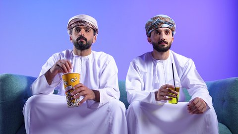 انشطة عطلة نهاية الأسبوع , بورترية لصديقان عربيان خليجيان عمانيان يرتديان الدشداشة و العمامة يستمتعان بوقت الفراغ , الاستمتاع بتناول الفشار أثناء مشاهدة الفلم , إيماءات وجه تدل على السعادة والاندماج بمشاهدة الأفلام والمسلسلات التلفزيونية, قضاء وقت ممتع في المنزل، تناول المشروبات المفضلة، خلفية زرقاء