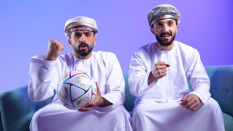 الحماس والتفاعل مع المنتخب، إيماءات الشغف والتشجيع للفريق، بورتريه لرجل عربي خليجي عماني يرتدي الدشداشة و العمامة يجلس على الاريكة يقوم بمشاهدة التلفاز و يحمل بيده كرة القدم برفقه صديقة، أنديه رياضية مختلفة، خلفية زرقاء