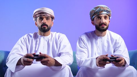 الاستمتاع باللعب في العاب الفيديو في اوقات الفراغ، مفهوم التسلية والترفيه و الفوز والانتصار، بورترية لرجلان عربيان خليجيان عمانيان يرتديان الدشداشة و العمامة يجلسان على الاريكة يحملان عصا التحكم، التحدي والمغامرة والتنافس، تقنيات حديثة متطورة، خلفية زرقاء