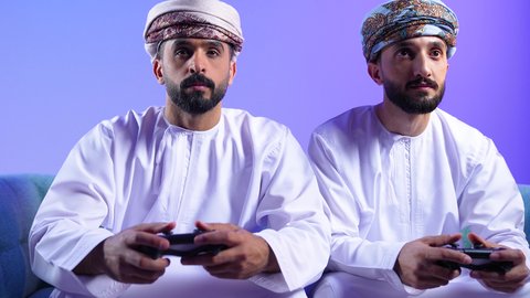 التحدي والمغامرة والتنافس، الاستمتاع باللعب في العاب الفيديو في اوقات الفراغ، مفهوم التسلية والترفيه و الفوز والانتصار،بورترية لرجلان عربيان خليجيان عمانيان يرتديان الدشداشة و العمامة يجلسان على الاريكة يحملان عصا التحكم، تقنيات حديثة متطورة، خلفية زرقاء