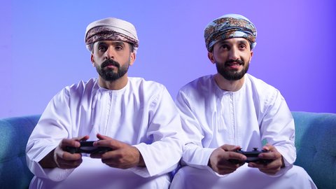 مفهوم التسلية والترفيه و الفوز والانتصار ، بورترية لرجلان عربيان خليجيان عمانيان يرتديان الدشداشة و العمامة يجلسان على الاريكة يحملان عصا التحكم، الاستمتاع باللعب في العاب الفيديو في اوقات الفراغ، التحدي والمغامرة والتنافس، تقنيات حديثة متطورة، خلفية زرقاء