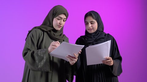 Follow-up and discussion of work, concept of preparing and reviewing scientific research, portrait of two Saudi Arabian Gulf women wearing the abaya holding white papers to review projects and work, proofreading work, reading notes, concept of culture and education, hobby of reading, colorful background