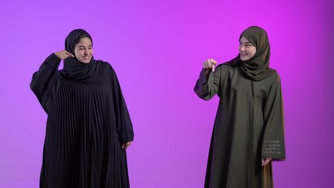 تحديد موقع شيء معين، إشارات وحركات يدين تدل على هذا المكان (هنا), بورتريه لامراتان عربيتان سعوديتان خليجيتان ترتديان الحجاب والعباءة يقومان بالاشارة بإصبع السبابة إلى شيء محدد, خلفية ملونه
