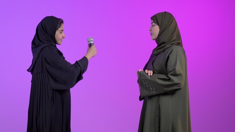 تقديم العروضات المسرحية، ايماءات وجه و يدين تدل على السعادة و الاستمتاع ، بورترية لامراه عربيه خليجيه سعودية ترتدي العباءه و الحجاب تحمل بيدها الميكروفون برفقه صديقتها ، موهبة الغناء ، خلفية ملونه