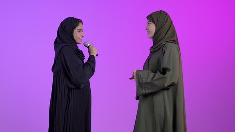 ايماءات وجه و يدين تدل على السعادة و الاستمتاع ، بورترية لامراه عربيه خليجيه سعودية ترتدي العباءه و الحجاب تحمل بيدها الميكروفون برفقه صديقتها ، موهبة الغناء ، خلفية ملونه
