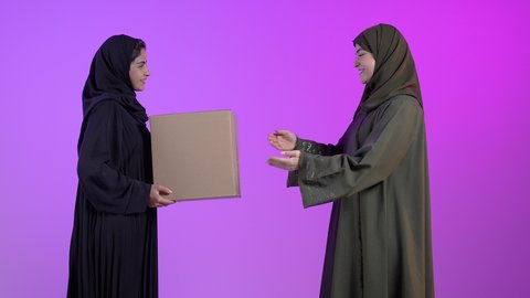 استلام الطلبات الالكترونية، بورتريه لامراتان عربيتان سعوديتان خليجيتان ترتديان الحجاب والعباءة تحملان صندوق مشتريات الاونلاين السريع، جودة شركات الشحن السعودية، خدمات التوصيل المنزل، خلفية ملونه
