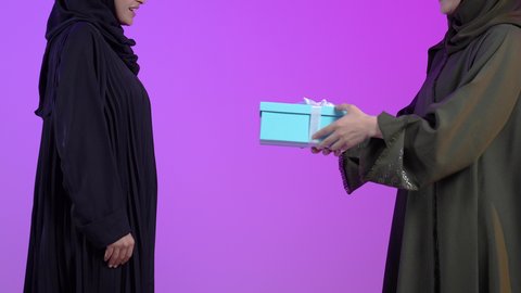 أعياد ومناسبات ومفاجآت سعيدة، مشاركة وتقديم الهدايا في الأعياد والمناسبات السعيدة، التعبير عن المحبة والألفة بين الاصدقاء, بورتريه مقرب ليد امراتان عربيتان سعوديتان خليجيتان ترتديان الحجاب والعباءة تقوم بتقديم الهدية لصديقتها, خلفية زرقاء