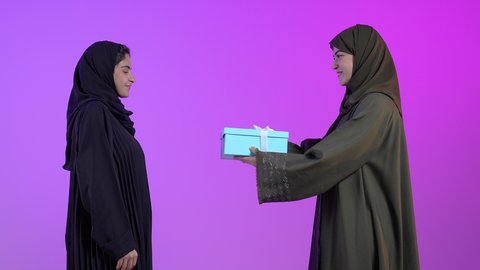 التعبير عن المحبة والألفة بين الاصدقاء، مشاركة وتقديم الهدايا في الأعياد والمناسبات السعيدة، أعياد ومناسبات ومفاجآت سعيدة, بورتريه لامراتان عربيتان سعوديتان خليجيتان ترتديان الحجاب والعباءة تقوم بتقديم الهدية لصديقتها, خلفية زرقاء
