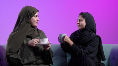 مشاركة المشروبات الساخن، بناء علاقات اجتماعية ناجحة، النقاشات والحوارات في الأجواء الهادئة، شابتان سعوديتان تقضي اوقات ممتعة، بورترية لإمرأة عربية خليجية سعودية ترتدي الحجاب والعباءة السوداء تجلس على الأريكة مع صديقتها بإيماءات السعادة، خلفية ملونه