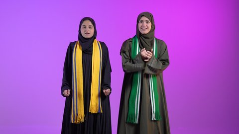 Integration, interaction and sports encouragement, having a good time watching the match, enjoying watching the national team, portrait of two Saudi Arabian Gulf women wearing the hijab and abaya raising their hands and raising the cup with gestures of happiness for victory and triumph, colorful background