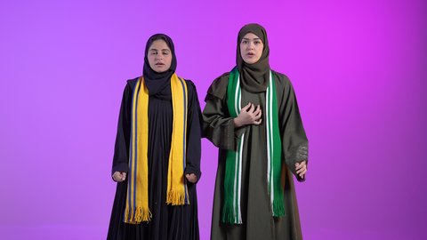 قضاء وقت ممتع بمتابعة المباراة، الاستمتاع في مشاهدة المنتخب، ، الاندماج والتفاعل والتشجيع الرياضي، بورتريه لامراتان عربيتان سعوديتان خليجيتان ترتديان الحجاب والعباءة يرفعان يديهما و ترفع الكأس بايماءات السعادة بالفوز والانتصار، خلفية ملونه