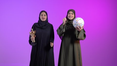 الاستمتاع في مشاهدة المنتخب، قضاء وقت الفراغ بمشاهدة المباريات، الاندماج والتفاعل والتشجيع الرياضي، ورتريه لامراتان عربيتان سعوديتان خليجيتان ترتديان الحجاب والعباءة يرفعان يديهما و ترفع الكأس بايماءات السعادة بالفوز والانتصار، خلفية ملونه