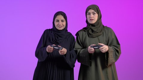 الاستمتاع باللعب في العاب الفيديو في اوقات الفراغ، مفهوم التسلية والترفيه و الفوز والانتصار ، بورتريه لامراتان عربيتان سعوديتان خليجيتان ترتديان الحجاب والعباءة يحملان عصا التحكم، التحدي والمغامرة والتنافس، تقنيات حديثة متطورة، خلفية ملونه