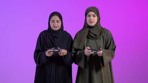 The concept of entertainment, fun, winning and triumph, a portrait of two Saudi Arabian Gulf women wearing hijab and abaya holding a joystick, enjoying playing video games in their spare time, challenge, adventure and competition, advanced modern technologies, colorful background