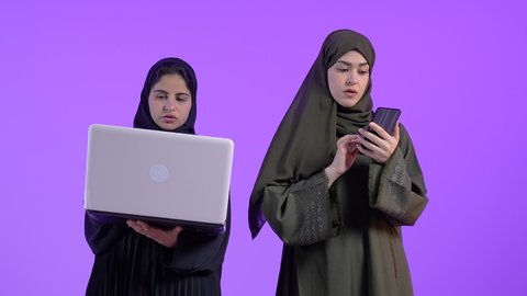 استخدام الأجهزة التقنية المتطورة,، دور التقنيات الحديثة في عملية البحث عن المعلومات المفيدة, بورتريه لامراتان عربيتان سعوديتان خليجيتان ترتديان الحجاب والعباءة احداهما تمسك بيدها الحاسب المحمول والاخرى الهاتف الجوال, ايماءات الفرح والحماس, خلفية ملونه