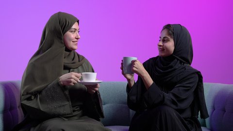 بناء علاقات اجتماعية ناجحة، النقاشات والحوارات في الأجواء الهادئة، شابتان سعوديتان تقضي اوقات ممتعة، بورترية لإمرأة عربية خليجية سعودية ترتدي الحجاب والعباءة السوداء تجلس على الأريكة مع صديقتها بإيماءات السعادة، مشاركة المشروبات الساخن، خلفية ملونه