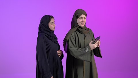استعمال الأجهزة التقنية الحديثة في اخذ صور تذكارية، بورتريه لشباتان عربيتان خليجيتان سعوديتان ترتديان العباءه و الحجاب يقومان بإلتقاط صورة سيلفي باستخدام الهاتف المحمول، ايماءات وتعابير البهجة والسرور، خلفية ملونه