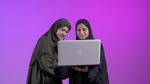 Browsing electronic applications and programs via laptop, using modern and advanced technical devices, remote business management, portrait of two young Saudi Arabian Gulf women wearing the abaya browsing the Internet via laptop with gestures of happiness and pleasure, colorful background