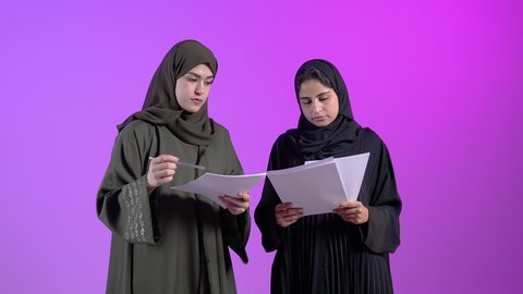 مفهوم التحضير ومراجعة البحوث العلمية ، بورتريه لشباتان عربيتان خليجيتان سعوديتان ترتديان العباءه يحملان بيدهما أوراق بيضاء للإستطلاع على المشاريع والأعمال ، تدقيق العمل ، متابعة ومناقشة العمل ، قراءة الملاحظات ، مفهوم الثقافة والتعليم ، هواية القراءة، خلفية ملونه