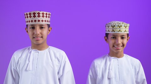 الوقوف باستقامة وثقه بالنفس، الاهتمام بالمظهر الخارجي،  بورترية لطفلان توأمان عربيان خليجيان عمانيان يرتديان الدشداشة و الكمه ينظران الى الكاميرا بايماءات السعادة، خلفية بنفسجية
